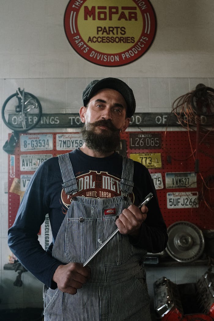 Bearded mechanic in overalls holding wrench in a garage adorned with license plates and vintage decor.